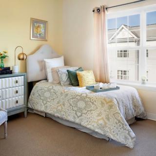 A cozy bedroom with a single bed adorned with cushions, a window allowing natural light, and tasteful decor.