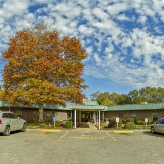 A small building with a parking lot and cars, surrounded by trees with autumn foliage under a partly cloudy sky.