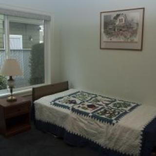 A cozy bedroom with a single bed adorned with a quilt, next to a nightstand and lamp by the window.