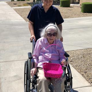 An elderly woman smiling in a wheelchair, accompanied by a caregiver on a sunny day.