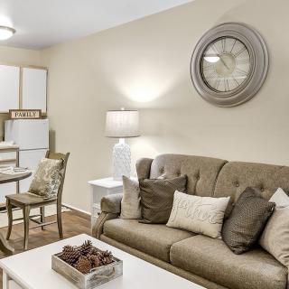 A cozy living room with a neutral color palette featuring a sofa, decorative pillows, and a large wall clock. A dining area is visible in the background.