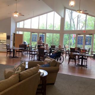 A spacious dining area with large windows, tables, chairs, and sofas, featuring ceiling fans and people seated, including one person in a wheelchair.