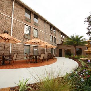A brick building with outdoor seating and umbrellas, surrounded by landscaped gardens and a fountain.