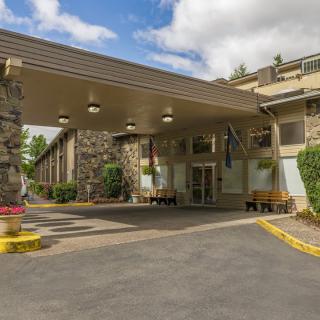 The entrance of a hotel with a wide canopy and stone accents, surrounded by landscaped greenery on a clear day.