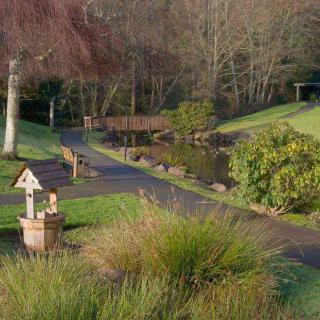 A scenic garden path with a bridge over a pond, surrounded by lush greenery and trees.