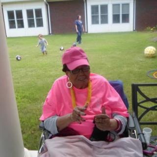 A person in a pink outfit sits in a wheelchair on a lawn, while children play in the background.