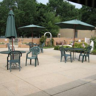 An outdoor patio with green chairs and tables, shaded by umbrellas in a garden setting.