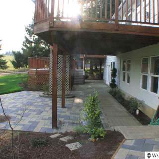 A backyard view featuring a patio area with a deck overhead, surrounded by greenery and landscaping.