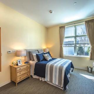 A cozy bedroom featuring a neatly made bed with striped bedding, a wooden nightstand with a lamp, and a large window with curtains letting in natural light.