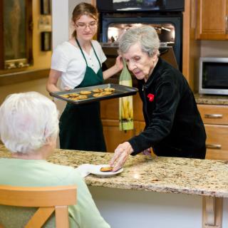 Two women baking cookies in a kitchen, with one serving an elderly person.