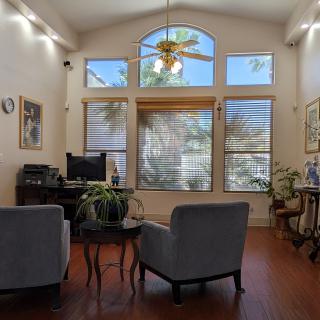 A cozy living room with large windows, featuring comfortable chairs, plants, and a ceiling fan.