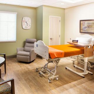 A well-lit hospital room featuring a patient bed with orange bedding and a comfortable seating area.