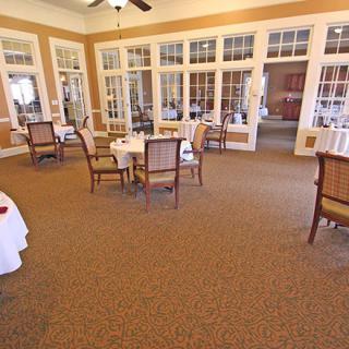 A bright dining room with tables and chairs set on a carpeted floor surrounded by large windows.