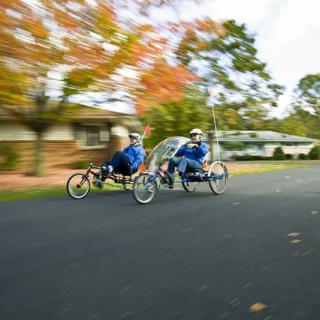 Two people riding recumbent bicycles down a tree-lined street in autumn.
