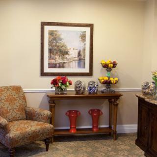 A cozy reception area featuring an armchair, a console table adorned with flowers and decorative items, and a framed painting on the wall.