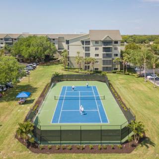 Aerial view of a tennis court surrounded by an apartment complex and greenery on a sunny day.
