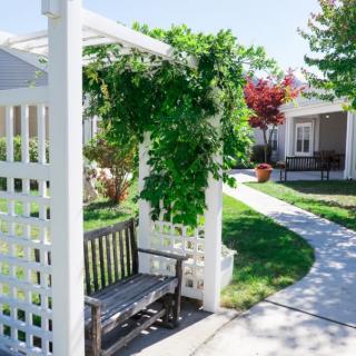 A picturesque garden scene featuring a white pergola adorned with greenery, a wooden bench, and a winding pathway leading to nearby houses.