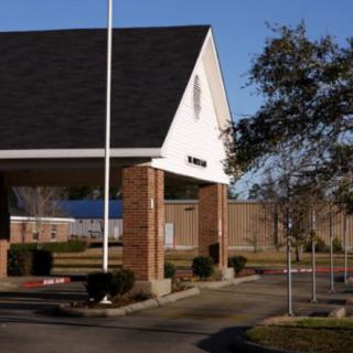 A small building with a steep roof and brick columns next to a driveway, featuring a flagpole and tree under a clear sky.