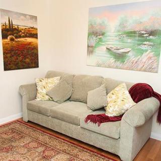 A cozy living room corner featuring a comfortable sofa adorned with cushions, flanked by two large paintings on the walls and a patterned rug on the floor.
