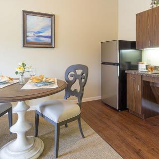 A cozy dining area with a round table, two chairs, and a compact kitchen setup featuring a refrigerator and wooden cabinets.