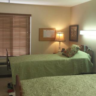 A cozy bedroom featuring two twin beds dressed in green linens, complemented by warm wooden accents and natural light filtering through the blinds.