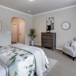 A modern, well-lit bedroom featuring a bed with a floral-patterned comforter, a gray armchair, a tall dresser, and tasteful wall art.