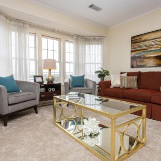 A cozy living room with a brown sofa, two gray armchairs, and a glass coffee table adorned with decor. Large windows bring in natural light, and a piece of artwork adds color to the walls.