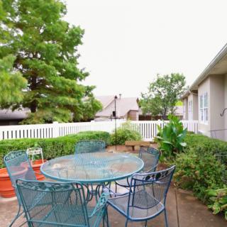 A charming patio area featuring a metal table and chairs, surrounded by lush greenery and enclosed by a white fence.
