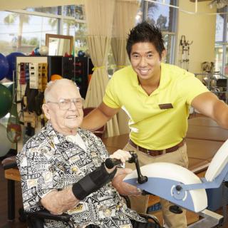 A healthcare professional assists an elderly person during a physical therapy session with exercise equipment.