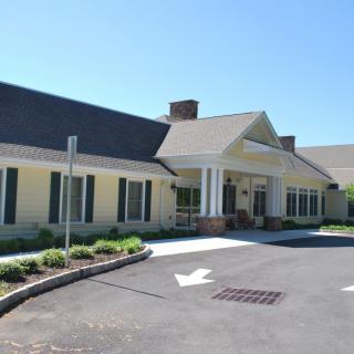 A single-story building with a sloping roof and large windows, surrounded by greenery on a sunny day.