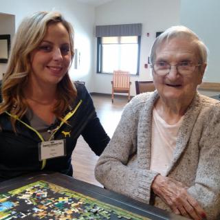 A caregiver sits next to a smiling elderly woman in an indoor setting with a puzzle on the table.