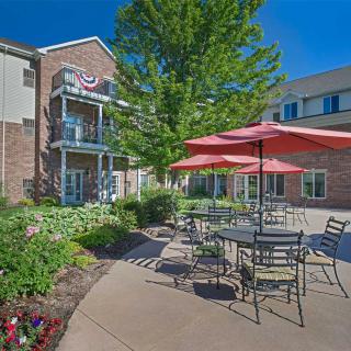 A well-landscaped patio area with tables, chairs, and red umbrellas set against a brick building on a sunny day.