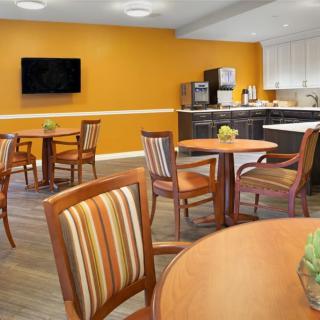 A cozy dining area featuring wooden tables and chairs with a striped design, an orange accent wall, a television, and a kitchenette.