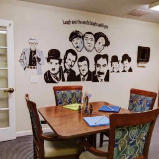 A decorated dining area with wall art featuring comedy legends above a wooden table set for meals.