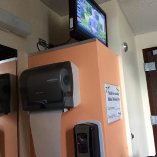 A bathroom corner featuring a hand dryer, a wall-mounted TV, and a dispenser.