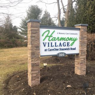 Entrance sign for Harmony Village at CareOne Stanwick Road surrounded by outdoor greenery.