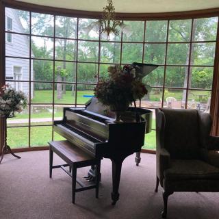 A grand piano sits in a bright living room with large windows overlooking a garden.