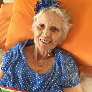An elderly woman is smiling while sitting in bed, wearing a blue polka dot dress and a festive birthday hat.