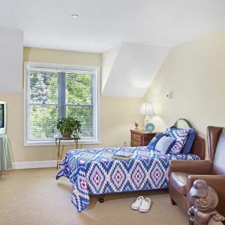 A cozy bedroom featuring a single bed, armchair, and television with natural light pouring in through a large window.