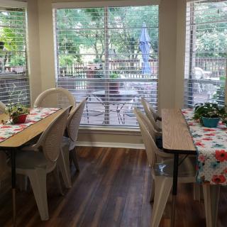 A cozy dining area with tables and chairs, adorned with plants and tablecloths, overlooking a garden through large windows.
