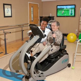 An elderly person engages in physical therapy with the assistance of a caregiver using fitness equipment.