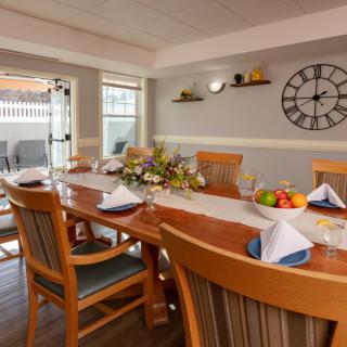 A cozy dining room with a wooden table set for a meal, featuring a decorative clock and shelves on the wall.