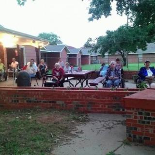 A group of people gathered on a patio in the evening, surrounded by trees and brick walls.