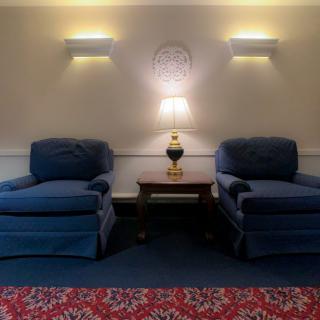 A cozy interior scene with two blue armchairs flanking a wooden table and lamp, set against a decorated wall with soft lighting.