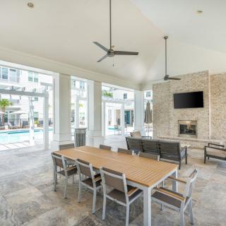A modern indoor dining area with a wooden table, chairs, and ceiling fans, featuring a fireplace and television, with large windows overlooking a pool.