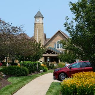 A picturesque building with a tower surrounded by lush gardens and yellow flowers, with a red car parked on the driveway.
