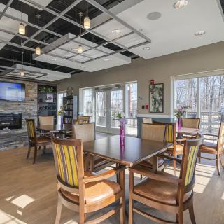 A modern dining area with tables, chairs, a television, and a fireplace, featuring large windows that let in natural light.