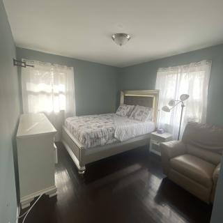 A cozy bedroom with a bed, armchair, and dresser, featuring two windows with sheer curtains.