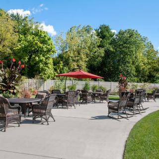 A sunny outdoor patio with tables, chairs, and a red umbrella surrounded by lush greenery.