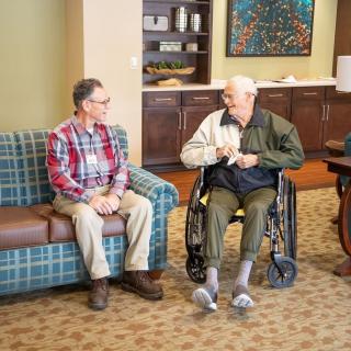Two elderly men chatting in a comfortable living room setting, one seated on a couch and the other in a wheelchair.